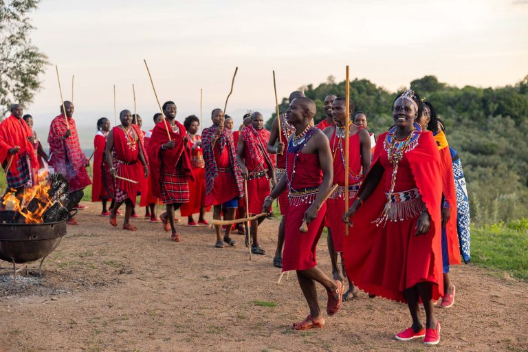 A Night Dance on the Savannah
The red shúkà cloth moves in sharp, rhythmic motions, glowing almost ember‑bright in the firelight.
Beaded necklaces and bracelets catch the light with quick flashes as the dancers jump in unison.
The bonfire crackle