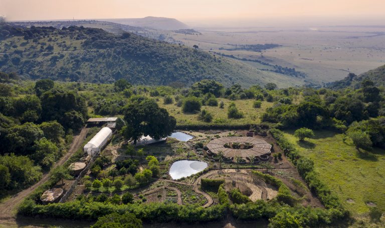 Aerial view of a lush green landscape with a Maasai Mara Manyata building structures, surrounded by trees and hills.